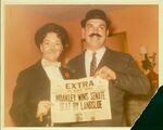 John Joseph Moakley and Evelyn Moakley, wearing suits and fake mustaches, holding newspaper with headline "John Joseph Moakley Wins Senate Seat By Landslide," 1960s by unknown